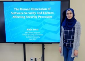 Hala standing next to a large TV with her title slide from her PhD proposal presentation