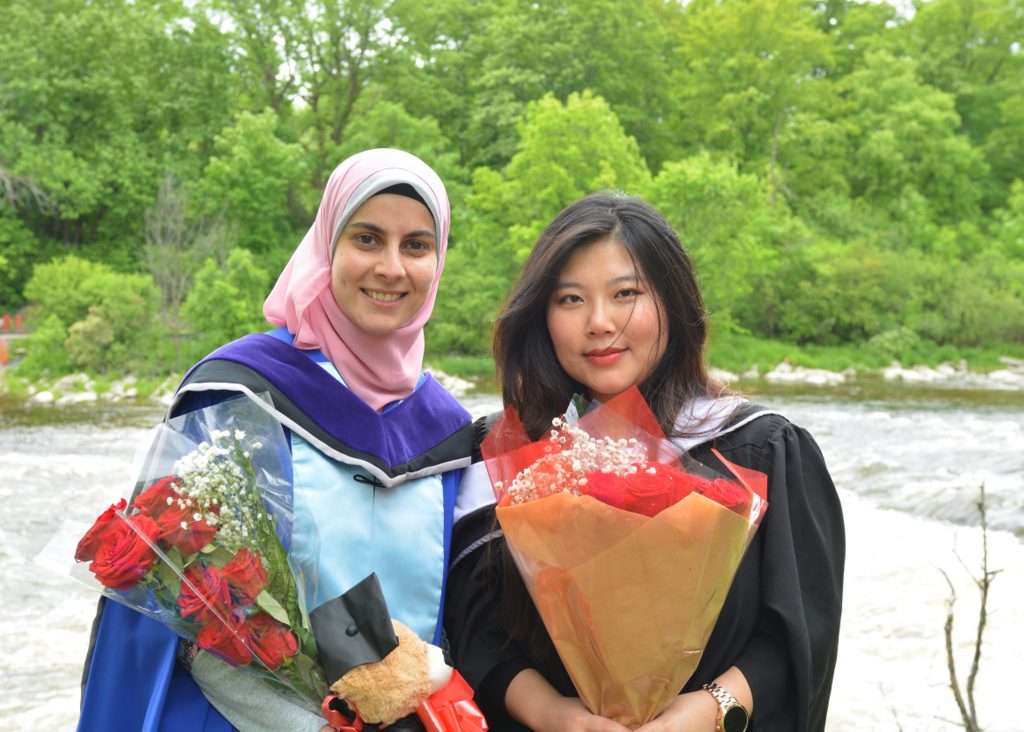 Hala and Becky standing by the river in their graduation gowns after convocation.