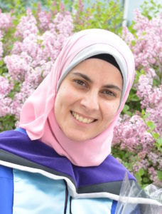 Headshot of Hala in her graduation gown, standing in front of a pink flowering shrub