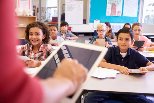 Children sitting in a classroom with their teacher; everyone holding tablet computers; Image credit:  Shutterstock Image number 388659994