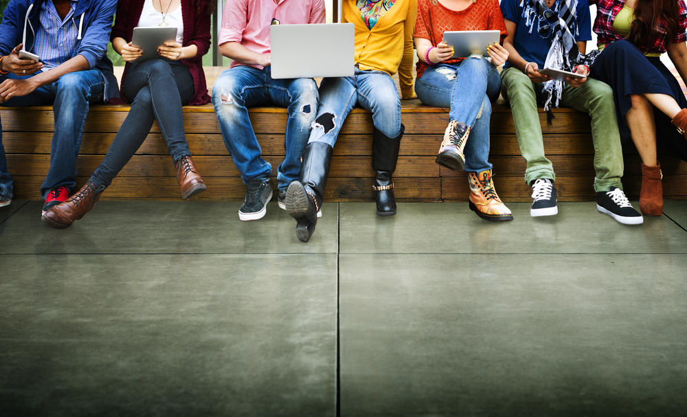 photo of seven youth sitting in line on a bench, wearing colourful clothing, and each holding some type of technology (tablet, phone, laptop).  The photo is cropped to show everyone from the shoulders down only. 