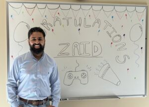 Zaid standing in front of a whiteboard in the lab that says "Congratulations Zaid"