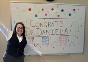 Daniela is smiling and standing in front of whiteboard that says Congratulations Daniela!